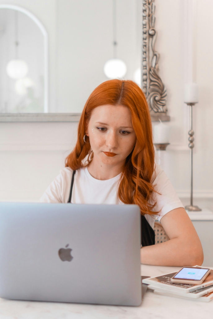 Redhead woman working on a laptop at a desk with a smartphone indoors.
